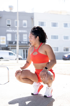 Woman in orange athletic outfit squatting outdoors with buildings in the background