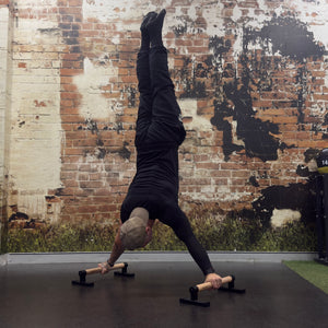 A man in a handstand position on a black gym floor, using a pair of wooden and metal parallettes. He is wearing a black t-shirt and black pants, set against a rustic brick wall background.