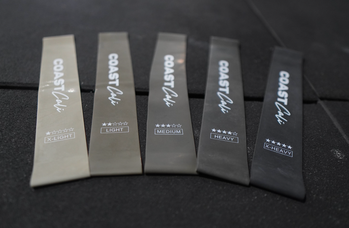 A set of grey mini resistance bands with varying resistance levels, labeled with the brand name 'COAST Cali', along with a black carrying pouch on a black gym floor