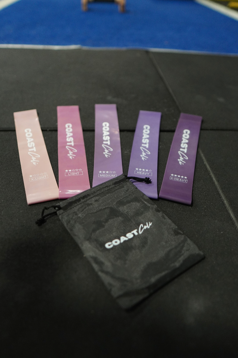 A set of purple mini resistance bands with varying resistance levels, labeled with the brand name 'COAST Cali', along with a black carrying pouch on a black gym floor