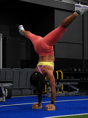 A woman in a handstand pose wearing a coral pink sports bra and leggings with a yellow waistband, performing a handstand on wooden blocks in a gym.