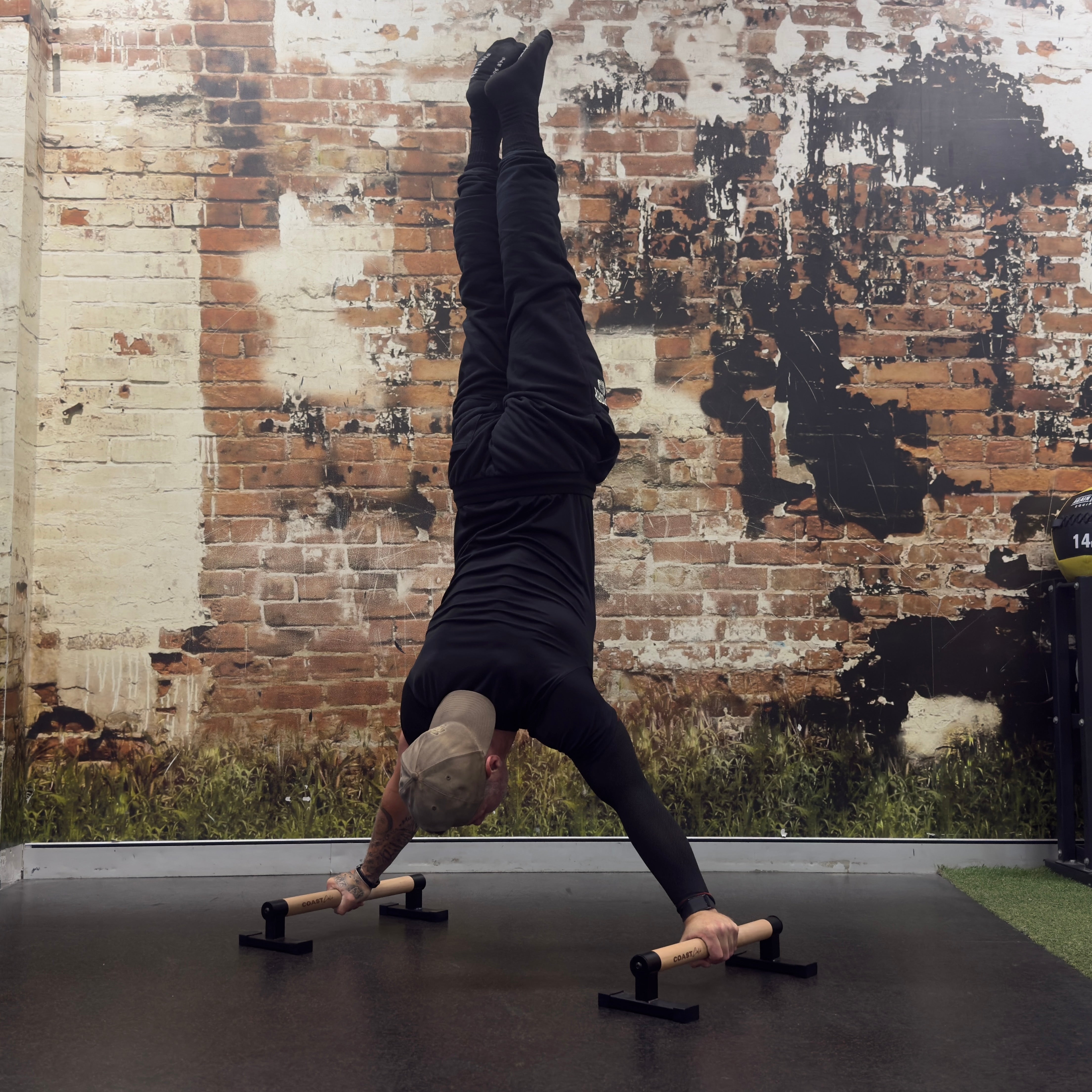 A man in a handstand position on a black gym floor, using a pair of wooden and metal parallettes. He is wearing a black t-shirt and black pants, set against a rustic brick wall background.