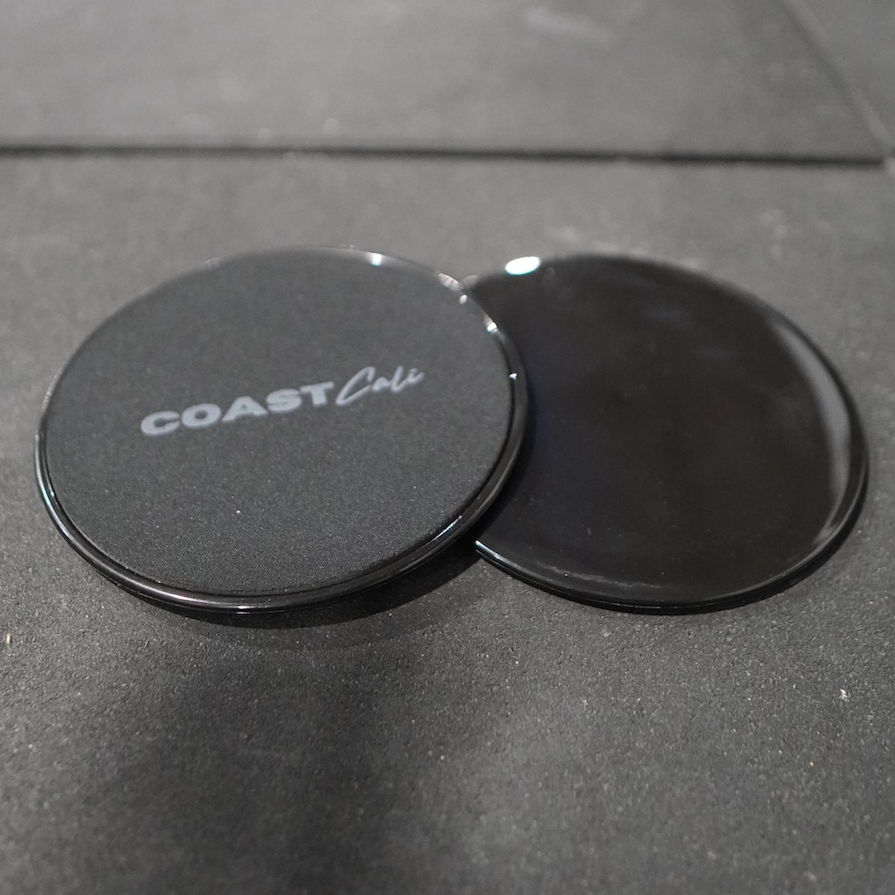 Two black gymnastics sliders with the logo 'COAST Cali' on the top surface in gym
