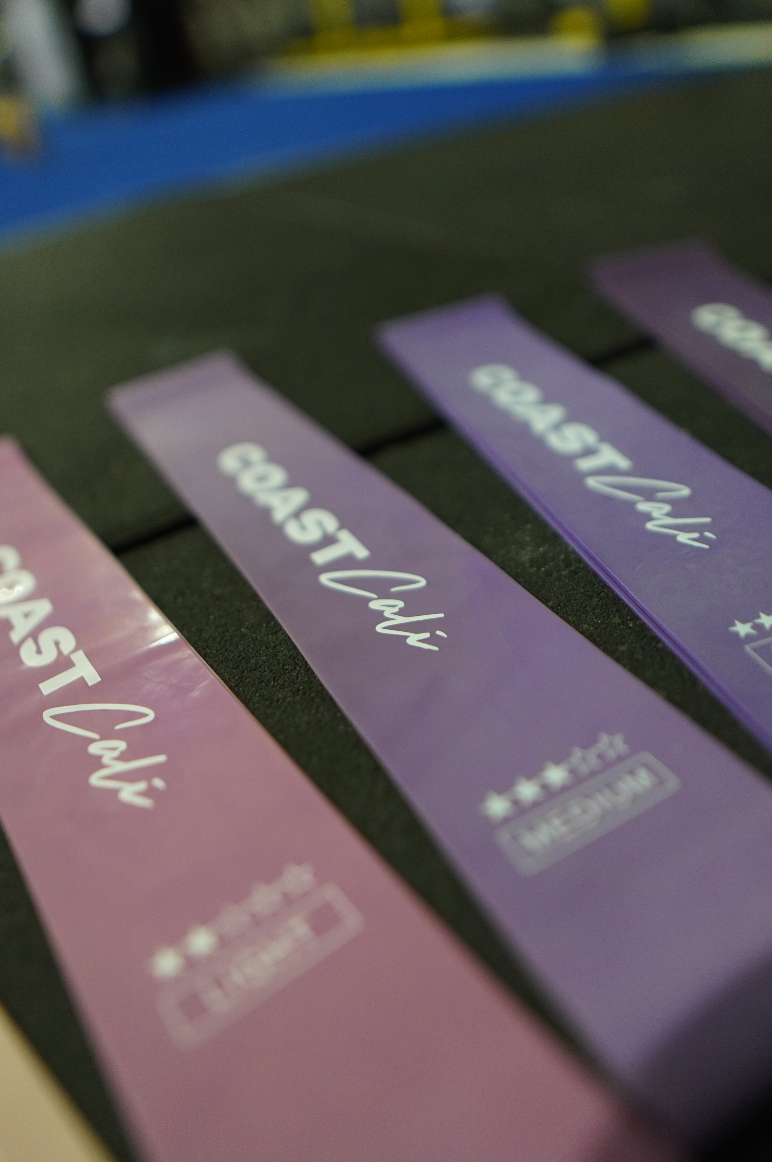 A set of purple mini resistance bands with varying resistance levels, labeled with the brand name 'COAST Cali', along with a black carrying pouch on a black gym floor