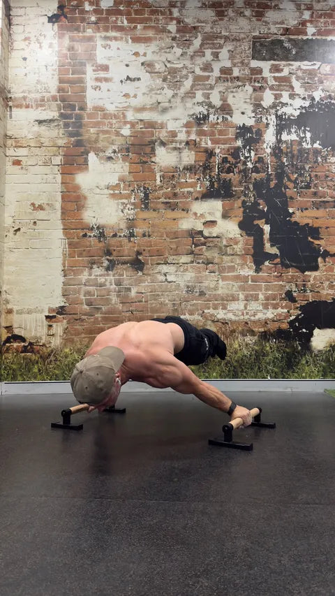 A shirtless man in a cap performs a plance on a pair of black parallettes on a dark gym floor, with a large brick wall mural in the background.